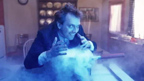 Rik Mayall is pictured under blue light while he smiles over a smoky saucepan in a dated kitchen with display plates behind him.