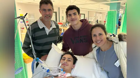 Jelenyela Pena Seb on a hospital bed has a hospital gown on, short black hair and a tube on his nose and throat. His mother is on his left, brother is above him, and his father on his left. The image has been taken on a hospital ward.