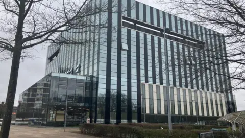Nadia Lincoln/LDRS A cube-shaped building with mirrored windows and landscaping and paving in front of the building.