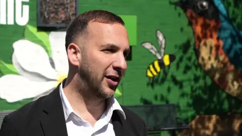 A man in a black blazer and white shirt looks towards a camera while standing in front of a dark green painted wall. 