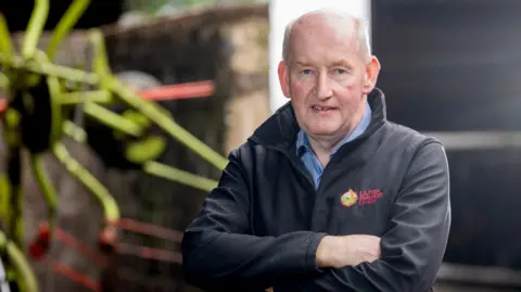 Glenn Cuddy is bald and has his arms folded. He's wearing a black jacket that says Ulster Farmers' Union in red lettering on the left breast. Under the jacket he is wearing a blue checked shirt.
