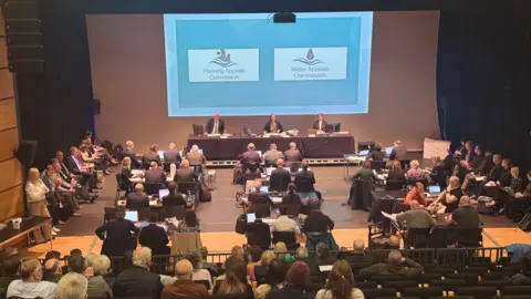 A three-person panel sits in front of a large screen in a theatre while lawyers and members of the public sit in front of them. The screen has captions on it reading "planning appeals commission" and "water appeals commission". The background is blue.