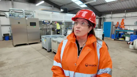 Nicola Henshaw is standing in a room where nitrous oxide canisters are being treated. She is wearing a high-vis jacket, with a range of machines behind her in the background.