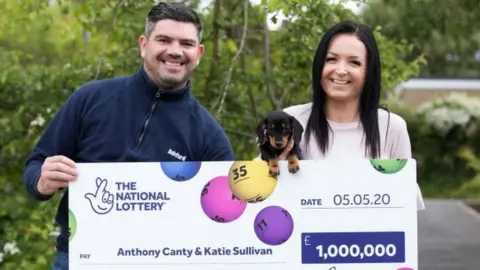 Anthony Canty is wearing a navy quarter zip jumper. He has dark hair and is holding a large cheque for £1m outdoors. Also holding the cheque, and a small black dog, is his partner, Katie Sullivan. She has long dark hair. Both are smiling.