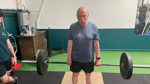 John Devine/BBC A man holds a weight lifting bar just below his waist with 50 kg on it, a trainer stands to the side giving instructions.