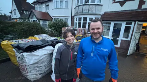 The boy in a grey top is next to his father, who is wearing a blue top and smiling at the camera. The man has dark hair and a beard and they are standing in front of a property.