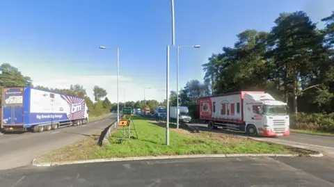 The view from a roundabout. It's a Google streetview image. A blue B&M roundabout is driving away on the lefthand side, and a red and white lorry is driving towards the camera on the right.