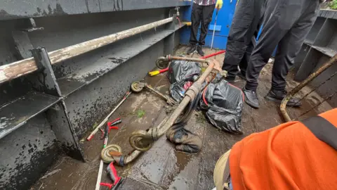 An old shopping trolley was dragged from the water. It is pictured on a boat with people standing around it. 