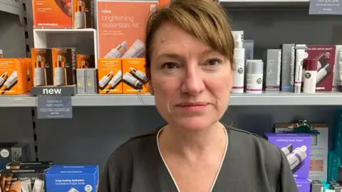 A woman with brown hair stands in her shop in the foreground, with skincare products on shelves in the background.