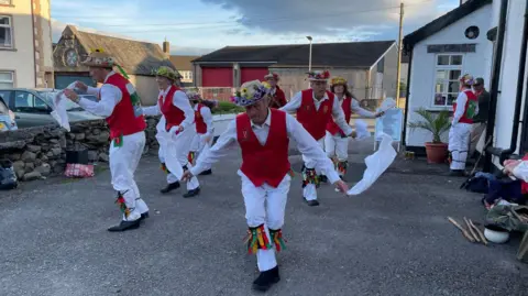 Members of Furness Morris Dance dancing in red and white clothing in Ulverston in May 2022. there are about eight of them wearing hats with colourful artificial flowers at the brim. 