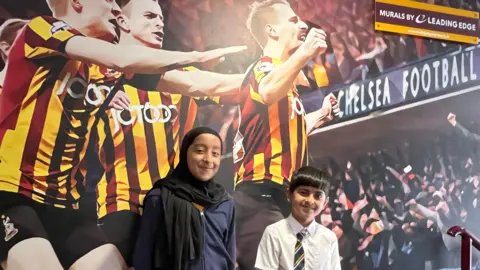 Aisha Iqbal/BBC Two young children standing in front of a poster wall which shows footballers celebrating. The girl wears a black headscarf and a zip-up jacket. The boy wears a white shirt and striped school tie.