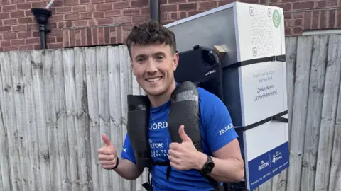 FTD Brothers Jordan wearing a blue t-shirt with a large white fridge mounted to his back. He is stood with with both thumbs up and he is smiling.