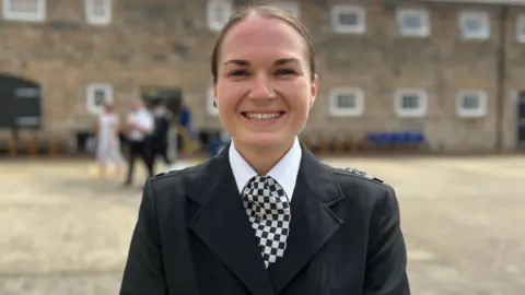 Jade Watkins in a black formal uniform with a checkered tie, standing in front of a stone building with blurred figures in the background.

