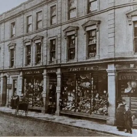 A black and white image of a Victorian-era shopfront with the name 'Fancy Fair' above the window