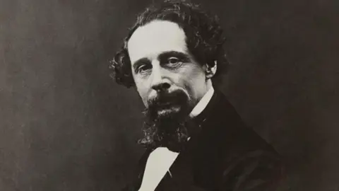 Charles Dickens Museum A black and white image of Charles Dickens he has curly balding hair and a goatee beared. He wears a black bow-tie and suit jacket.