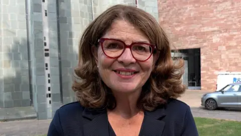 Diana Johnson looks at the camera. She is wearing a navy blouse and red eyeglasses. She has brown shoulder length hair and is standing in front of Coventry Cathedral.