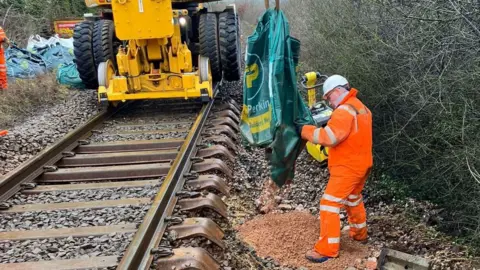 Network Rail A rail engineer in orange high-vis overalls shakes out a Travis Perkins bag containing building material in it onto a train line. A large machine is on the tracks.