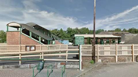 General view of a train station. A cream picket fence divides the road from the railway track area. Steps and a bridge have been built over the top of the track. 