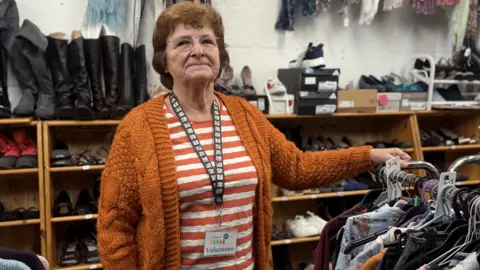 An older woman with light brown hair. She is wearing a stripped orange and white shirt and is wearing an orange cardigan. She is leaning on a clothes rack and has shoes and boots in the background