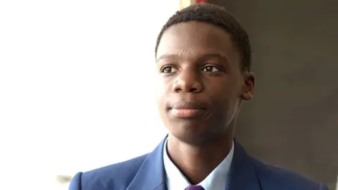 Shaun Whitmore/BBC A head and shoulders image of a young man. He is wearing a navy blazer over a smart shirt. He is looking away from the camera.