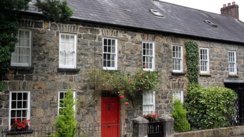 Grey stone terraced houses with green and red plants. The front door is red and the windowframes white.