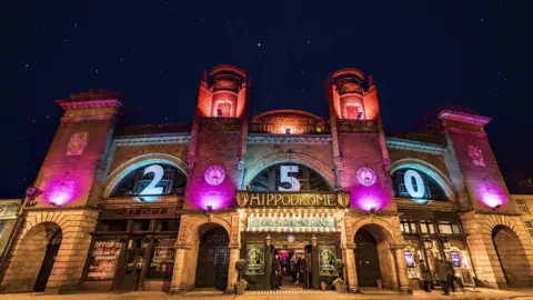 Great Yarmouth Hippodrome A view of the outside of the Great Yarmouth Hippodrome at night. The sandstone coloured bricks are floodlit by purple lights.