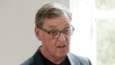 A man with grey hair and glasses is speaking at a podium, which is not in frame. He is wearing a grey jacket and dark shirt