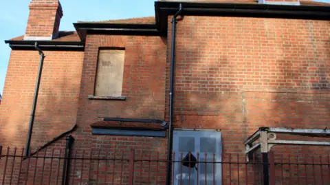 A general view of a dilapidated house. It is a red-brick home with a window boarded up with wooden board.