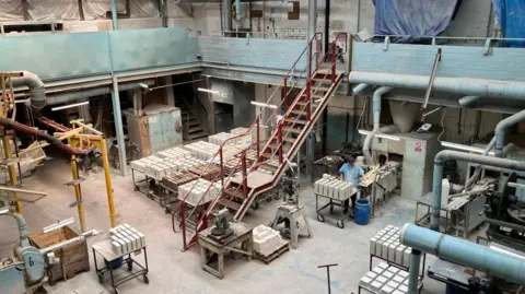 A large two-storey industrial space, with large metal steps leading to the ground floor, where tiles are stacked in blocks on wheeled tables