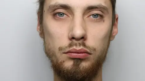 A man with short brown hair and brown or ginger facial hair and blue eyes. It is a custody photo and the background is grey.