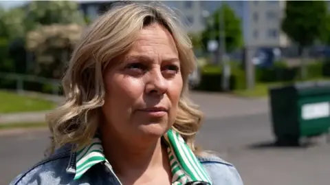 Katriona has collar length blonde hair. She is wearing a green and white striped shirt under a light blue denim jacket