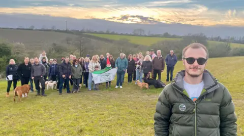 A man in sunglasses, a green puffer jacket and grey t-shirt is standing in a field which dips into a valley. Behind him are thirty adults, six children and eight dogs. Four members of the group is hold a banner which reads ‘Save S12 Green Belt’. In the distance are two fields with a hedge boundary coming up out of the valley. There are trees and a school on the edge of the fields. The sun is setting.