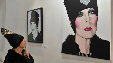 BBC A woman who is wearing a black dress and head wrap is stood in the left of the picture and is looking at a painting of herself in a black wrap with pink lipstick and pink blush. There is a second black and white, smaller painting of her to the left of that picture