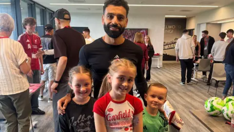 Family photo Mo Salah, with black hair, a black beard and a black long-sleeved Adidas training top, smiles at the camera with three young children - two girls and a boy - standing in front of him. 