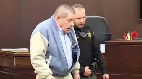 Reuters An older man in a light blue jacket is escorted in a courtroom by a law enforcement officer in a dark uniform