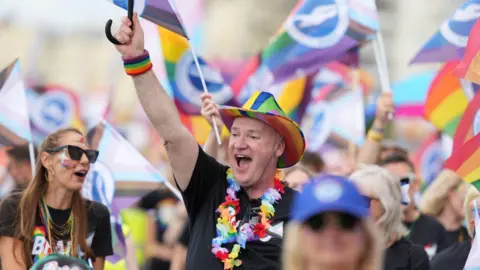 Eddie Mitchell A man with a rainbow hat on. He is smiling towards a crowd of people.