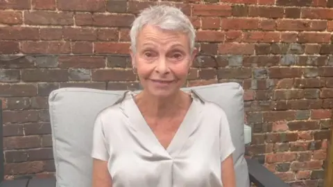 NHSBT Maggie Hackney pictured outdoors on a garden seat with a brick wall behind her. She smiles at the camera and wears a beige/grey silk short-sleeved shirt with a v-neck and gold hoop earrings. She has short pixie style grey hair.