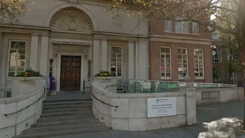 The exterior of the entrance to a council building. It is a brick and concrete structure with steps leading to a wooden door. The front of the building has thin windows.