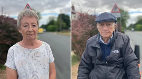 A woman wearing a white flowery top looks towards the camera with a stern look on her face next to a road. On the right, a man wearing a cap and jacket looks towards the camera while sat in his mobility scooter.