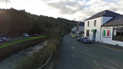 A river is on the left side of the picture, with a single lane carriageway running parallel to it. On the right of the picture there is a white rendered building, that appears to be an Italian restaurant with a green, white and red sign on the wall 