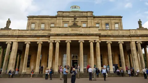 Pittville Pump Rooms - a honey coloured spa building in Cheltenham with lots of pillars - on a busy day, with a lot of older people stood outside