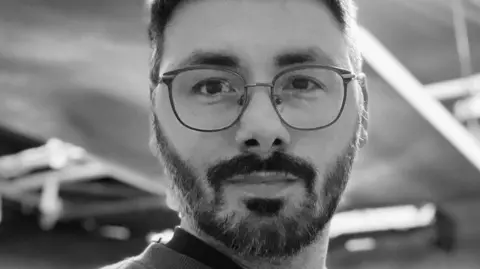 Number Nine PR A black-and-white photograph of Baz Sells who has short dark hair, a short dark beard and wears round glasses.