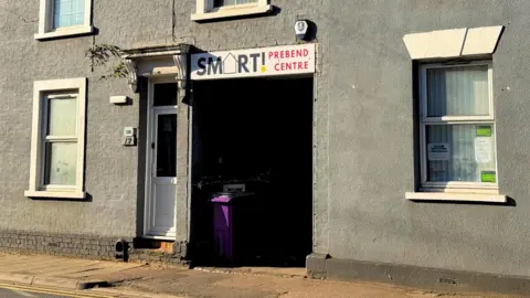 The outside of a day centre, showing a grey building with white windows, a purple rubbish bin, a sign above a gap and a path in front. An alarm is on the wall and a doorway is to the left, with a tree growing out of the top of a lintel.