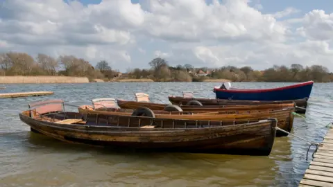 Boats moored on the water at Hornsea Mere. There are trees in the background and the sky is blue with fluffy cloud.