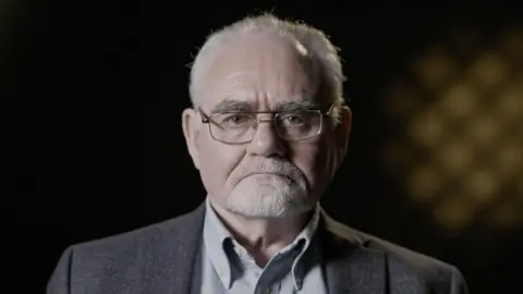  BBC/Keo Films Patrick Magee, a man with a grey shirt, grey jacket, glasses and short grey hair and a short grey beard.