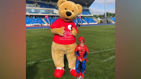 A young boy dressed in a blue and red Spider-Man costume with a matching mask over his face. Next to him is a person dressed as a large bear with a red t-short. Text on it in white reads "Birmingham Children's Hospital". The bear has red shoes with white laces