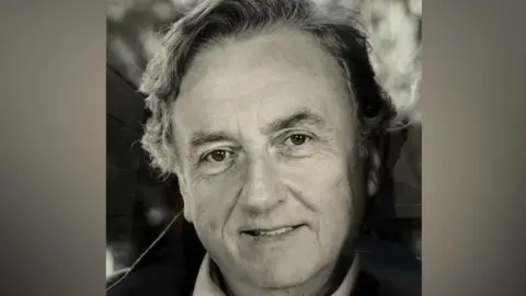 A black and white head and shoulders image of a man smiling at the camera. He has grey hair and is wearing a light-coloured shirt.