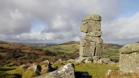 Dartmoor National Park Authority Nine giant boulders are piled on top of each other on a grassy outcrop. There are singular boulders lying on the ground nearby. All of them are covered in lichen. Dartmoor stretches out behind them, covered with fields, hedges and bracken. It is sunny but there are also clouds. 