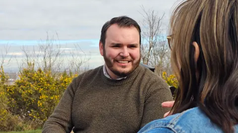 Sean Houlston smiling in a dark green jumper with an unknown woman in the foreground