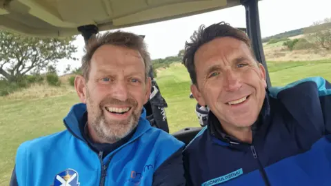 Supplied A selfie of Justin Middleton and David Scott who are wearing blue fleeces and sat in a golf cart.